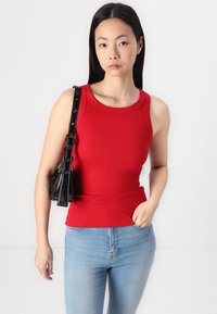 Camiseta de tirantes roja con cuello redondo, combinada con jeans de color azul claro. El modelo sostiene un bolso negro de hombro con herrajes en plata.