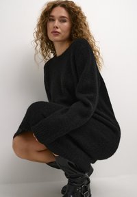 Juoda, tekstūruota mezginio suknelė su laisvu siluetu, ilgomis rankovėmis ir šoniniais sklendais, dėvima su juodais iki kelių batais su sagtimis.