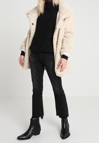 Manteau en polaire beige avec un grand col, porté sur un col roulé noir. Jeans courts noirs et bottines noires complètent la tenue.