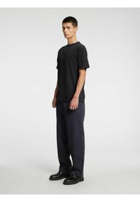 Melnas kokvilnas T-krekls ar brīvu piegriezumu, īsām piedurknēm un apaļu kakla izgriezumu, kopā ar vaļīgām tumši zila tonī biksēm un melniem apaviem.