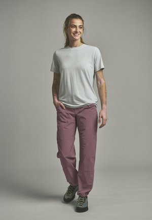 Young woman smiling, wearing light gray t-shirt, mauve pants, and gray hiking shoes, standing with one hand in pocket against gray background.