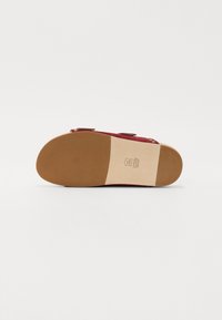 Semelle d'une sandale rouge avec une semelle extérieure en caoutchouc marron, une semelle intermédiaire en cuir beige, et des sangles à boucle, photographiée sur un fond blanc.