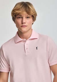 Lys rosa polo-shirt med krave, der har en tre-knaps placket og et lille mørkt logo af en golfspiller på venstre bryst. Blød tekstur.
