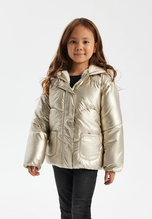 Metallic guld pufferjakke med en faux fur-fort opskrift, forlommer og knaplukning, båret over en mørk top og jeans.