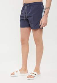 Pantaloni da nuoto blu navy con vita elastica e cordino, caratterizzati da due strisce verticali azzurro chiaro sui lati. Indossati con sandali bianchi a scivolo.