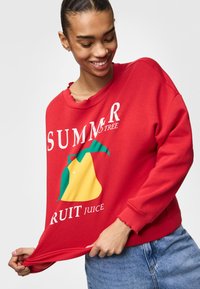 Sudadera roja con un gráfico de una pera amarilla y hojas verdes. El texto dice "JUGO DE FRUITA DEL ÁRBOL DE VERANO". Tela suave, corte relajado.