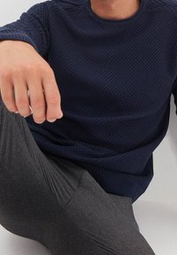 Muž v texturovaném námořnickém svetr a šedých kostkovaných kalhotách, sedící s předpaženou rukou proti bílému pozadí.