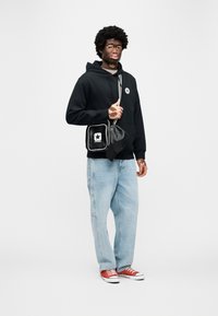 Μαύρο φούτερ με κ ουκούλα και λογότυπο, ανοιχτό μπλε τζιν παντελόνι, κόκκινα αθλητικά παπούτσια και μια μικρή μαύρη τσάντα με λευκό λογότυπο αστεριού και περιθώριο.