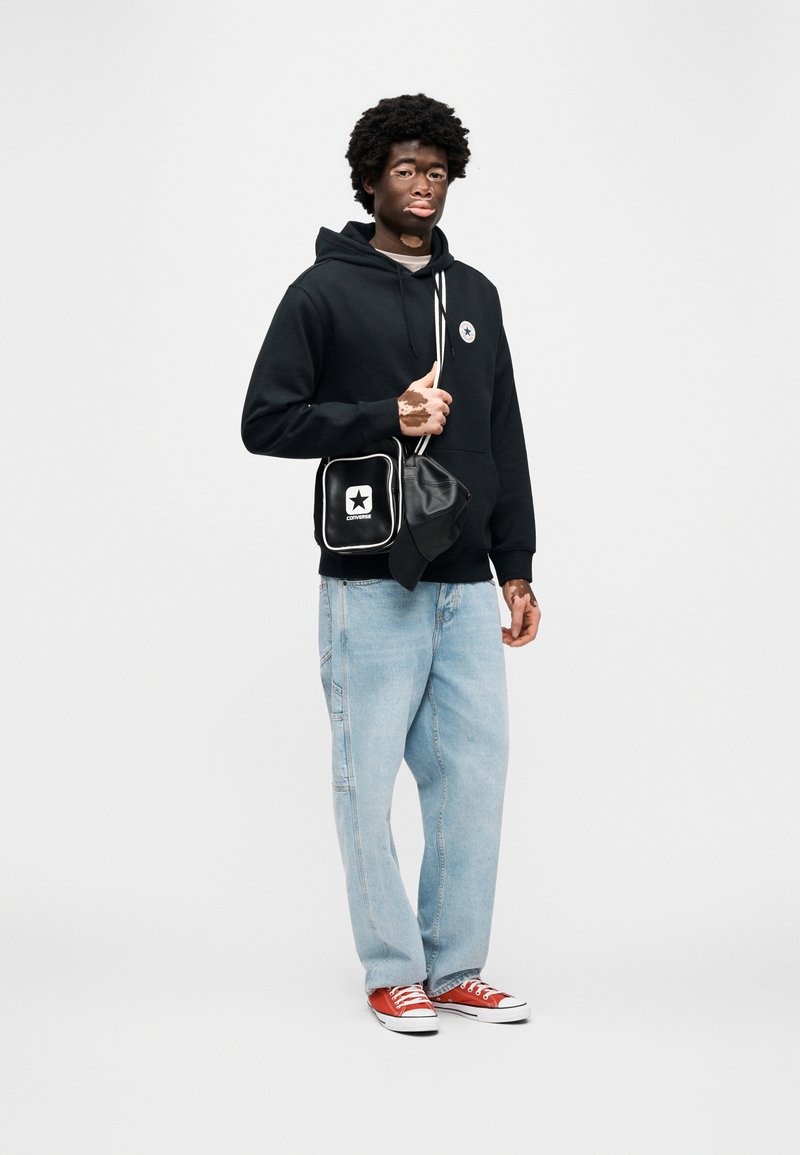 Sort hættetrøje med hætte og logo, lyseblå denimjeans, røde sneakers og en lille sort taske med hvidt stjerne-logo og kant.