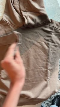 Světle hnědá mikina s kapucí z měkké tkaniny, která má klokaní kapsu, stahovací kapuci a strukturovaný povrch s viditelnými švy.
