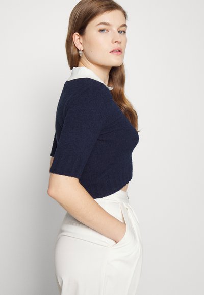 Sweater curto azul-marinho com um tricot texturizado, mangas curtas e um colarinho branco. O modelo usa calças creme com bolsos laterais.