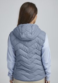 Vesta gri matlasată cu glugă și model în zig-zag. Purtați peste un top cu mânecă lungă de un albastru deschis, cu manșete și tiv texturate.