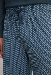Close-up van een persoon in blauw- en zwartgekruiste drawstringbroek en een donkere lange mouw shirt, hand rustend naast hun zij.