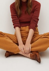 Henkilö istuu jalat ristissä, päällä rustinvärinen neule, sinapinkeltaiset housut ja ruskeat loaferit, kädet lepäilevät jaloilla tasaisella taustalla.