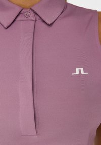 Ärm-lös polo skjorta i mjukt lavendelfärgat tyg med klassisk krage, knappstängning fram och en liten vit logotyp på vänster bröst.