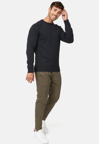 Suéter de punto gris oscuro con puños y dobladillo acanalados, combinado con pantalones oliva claros y zapatillas blancas, en un hombre que está de pie con una mano en la cabeza.