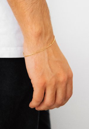 Guldkædearmbånd på et håndled, med sammenflettede led og en glat, poleret finish, mod en lys hudtone og ensfarvet baggrund.