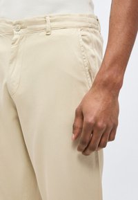 Άντρας που φοράει μπεζ παντελόνι και λευκό ριμπ πουκάμισο, κοντινό πλάνο του δεξιού χεριού που ακουμπάει δίπλα στο μηρό.