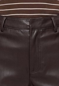 Pantalon en cuir marron foncé avec passants pour ceinture, couture avant visible, porté avec une chemise marron comportant des bandes horizontales blanches à la taille.
