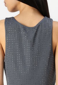 Mujer vistiendo una camiseta sin mangas de color gris oscuro con pequeños remaches plateados espaciados uniformemente en la parte posterior, mostrando piel y su largo cabello oscuro.