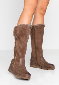 Brązowe zamszowe kozaki na koturnie sięgające za kolano z bocznymi zamkami, sztucznym futrem, detalem klamry i teksturowanymi gumowymi podeszwami noszone na gołych nogach.