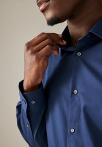 Camicia blu scuro a maniche lunghe con tessuto testurizzato. Presenta un colletto e bottoni argentati sul davanti e sui polsini.