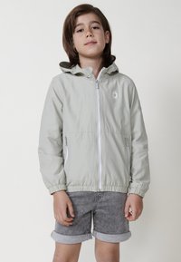Giacca leggera verde chiaro con cappuccio, chiusura con zip e polsini elasticizzati; abbinata a pantaloni corti in denim grigi arrotolati.