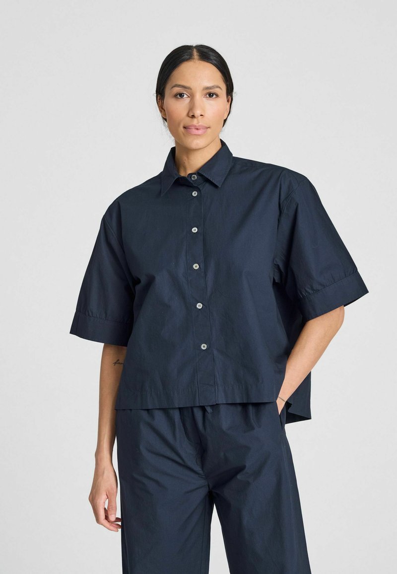 Camicia button-up blu navy a maniche corte con colletto, vestibilità comoda e texture liscia. Presenta bottoni chiari e un orlo corto.