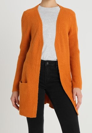 Femme portant un cardigan orange côtelé ouvert à l'avant avec des poches frontales, un t-shirt gris clair et un jean noir taille haute.
