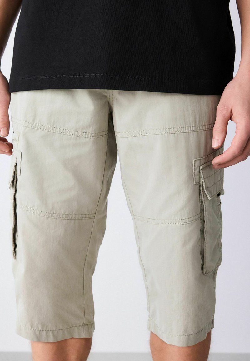 Beżowe krótkie spodnie cargo z klapkami noszone przez osobę w czarnej koszulce, ukazujące sylwetkę od pasa do nieco poniżej kolan.