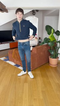 Sudadera de media cremallera en color navy con un logo, combinada con jeans ajustados azules y zapatillas blancas. Entorno interior con un sofá de pana marrón y plantas.