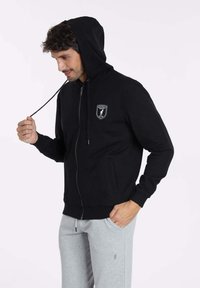 Veste à capuche zippée noire en tissu doux, dotée d'une poche avant, d'une capuche avec cordon de serrage réglable et d'un logo en badge sur la poitrine.