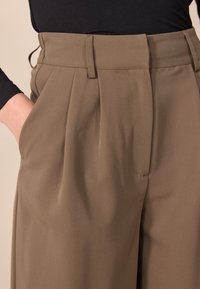 Hogewaisted, geplooide broeken van een effen taupe stof. Voorzien van zijzakken en een zachte textuur met zichtbare taillebanddetails.