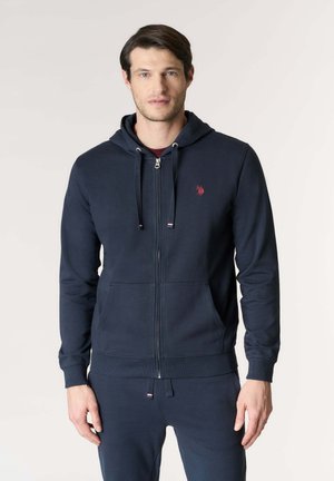 Uomo che indossa una felpa con cappuccio e cerniera blu navy con tasche frontali e pantaloni abbinati blu navy, in piedi contro uno sfondo chiaro uniforme.