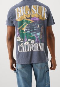 Graphic t-shirt featuring a blue-gray color with "BIG SUR CALIFORNIA" text and a landscape print of mountains, ocean, and flowers in vibrant colors.