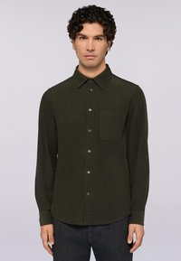 Camicia a maniche lunghe verde scuro con bottoni, in un tessuto texturizzato, dotata di colletto a punte e un'unica tasca sul petto. Presenta un orlo arrotondato e chiusura con bottoni.