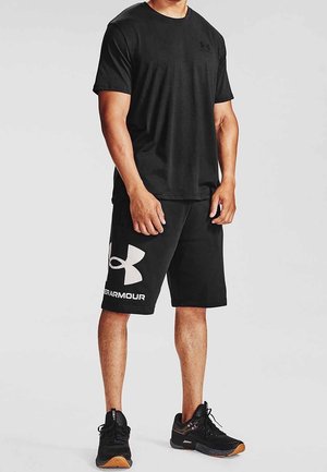 Homme portant un t-shirt noir Under Armour, un short noir avec un logo blanc et des chaussures de sport noires, debout sur un fond neutre.