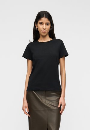 Hosszú sötét hajú nő, egyszerű fekete rövid ujjú pólót és sötétbarna bőrszoknyát visel, sima, világos háttér előtt áll.