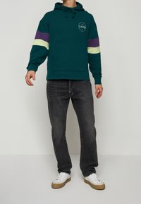 Sudadera color azul verdoso con rayas moradas y verdes limón en las mangas, bolsillo tipo canguro y detalle del logotipo. Combinada con jeans gris oscuro y zapatillas blancas.