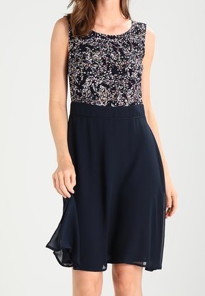 Mouwloze jurk met een bovenstuk met bloemendessin en een effen donkere, vloeiende rok, gedragen door een vrouw die de zoom van de rok vasthoudt.