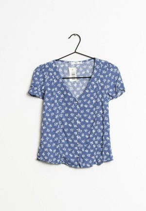 Blouse en tissu bleu à manches courtes. Présente un décolleté en V et un motif floral blanc. Fermeture à boutons sur le devant.