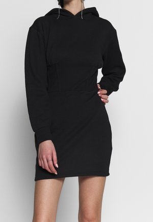 Femme portant une robe à capuche noire à manches longues avec une taille cintrée, une main sur la hanche, sur un fond blanc uni.