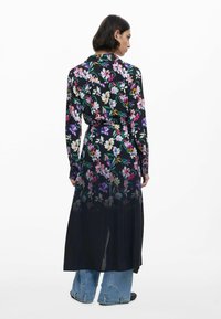 Blumen Kleid mit langen Ärmeln in Schwarz, das mit leuchtend bunten Blumen verziert ist, mit Taillierung und fließender Silhouette. Getragen mit blauen Jeans.