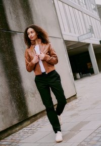Woman with curly hair, wearing a brown leather jacket, white shirt, black pants, and white sneakers, walking on a paved urban sidewalk.