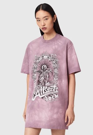 Jeune femme portant une robe T-shirt ample à effet tie-dye violet avec un graphique en noir et blanc représentant un squelette couronné de fleurs et le texte « AllSaints International Rock Festival ».