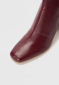 Stivale alla caviglia in pelle bordeaux con punta quadrata e texture liscia. Presenta un sottile dettaglio di cucitura lungo la parte superiore. Tacco a forma quadrata.