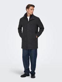 Dunkelgrauer Wollmischmantel mit Stehkragen, vorderen Knöpfen und zwei Seitentaschen. Kombiniert mit blauer Jeans und schwarzen Schuhen.