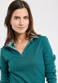 Polo a maniche lunghe color teal con colletto a stampa floreale, scollo a V e una texture liscia. Design semplice e aderente.