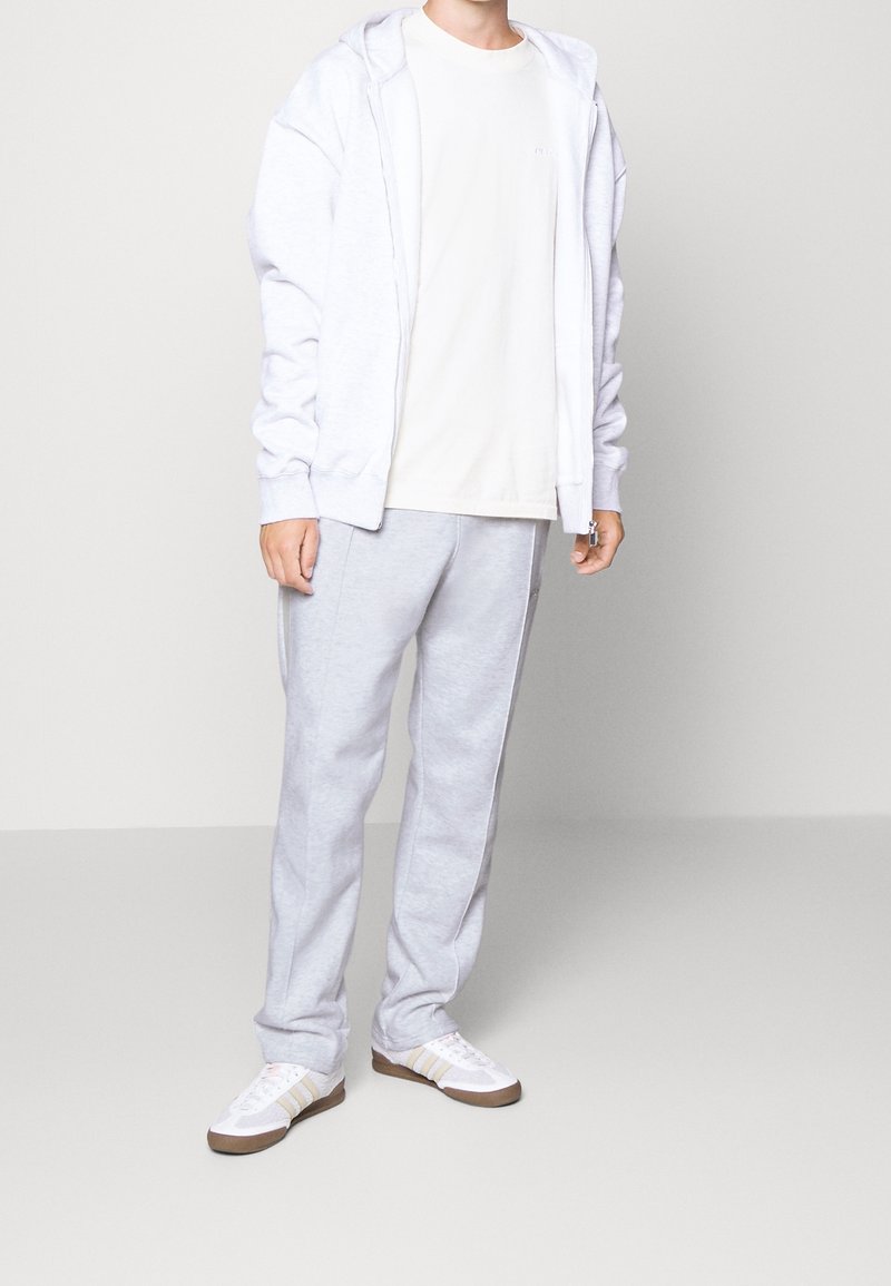 Světlá šedá mikina s kapucí a dlouhými rukávy, doplněná o světlé šedé tepláky. Bílá teniska s béžovými akcenty dokončuje celý outfit.
