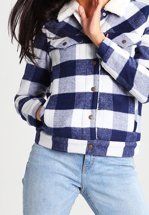 Personne portant une veste à carreaux bleue et blanche avec un col sherpa blanc et un jean bleu clair, main dans la poche et près du visage.